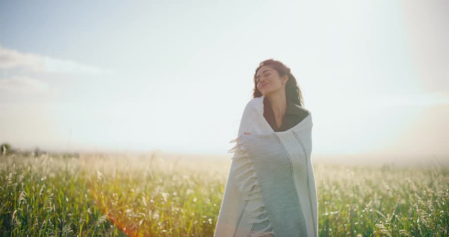 A serene young woman wrapped in a blanket enjoys the warm sunshine and gentle breeze while standing in a beautiful, sunlit field of tall grass