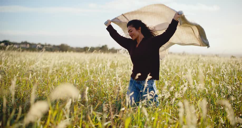 A happy young woman walks through a sunlit golden field with a blanket flowing in the wind