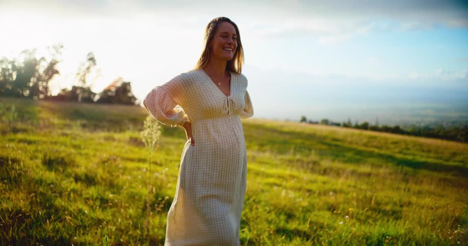 A beautiful pregnant woman smiles peacefully as she walks through a golden sunlit field, stopping to gently hold her belly