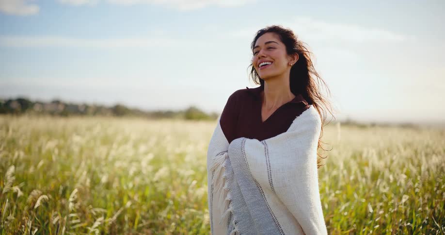 Portrait of a beautiful young woman wrapped in a warm blanket smiling peacefully in a sunlit field as the wind blows her hair