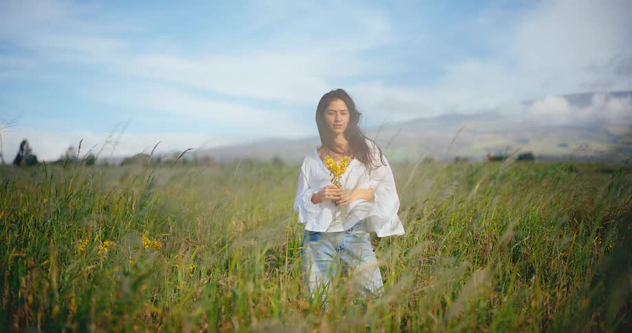 A serene young woman with yellow wildflowers enjoys the sun and wind blowing through her hair in a beautiful green field