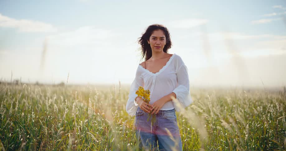 A beautiful young woman in a white blouse holds yellow wildflowers while standing peacefully in a sun-drenched field of tall grass