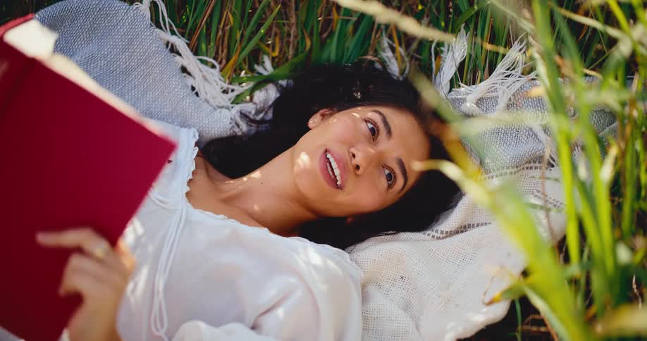 A serene woman with a bright smile lies on a blanket in a sunlit grassy field, holding a red book and basking in the warmth