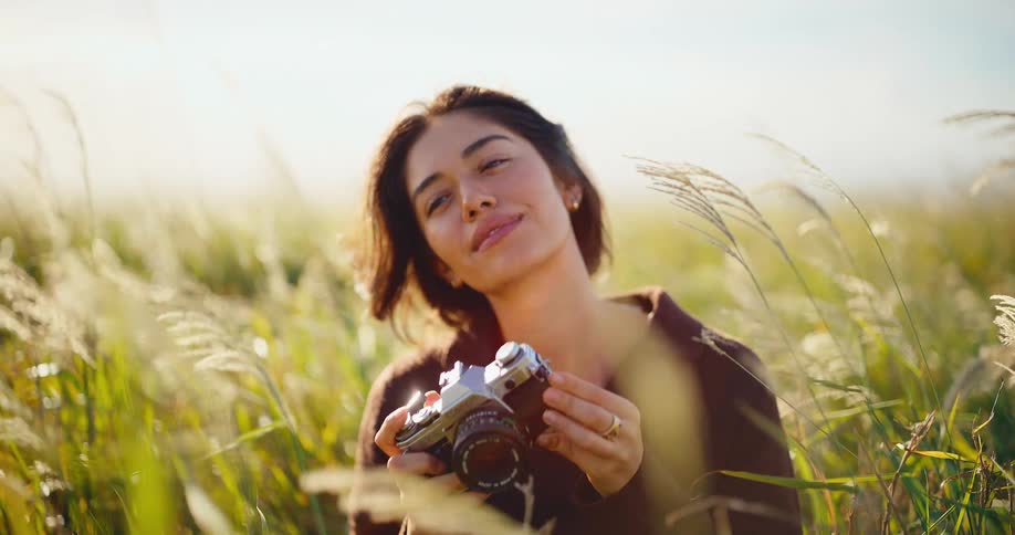 A serene young woman smiles after taking a photo with her vintage camera in a sun-drenched field of tall grass in slow motion