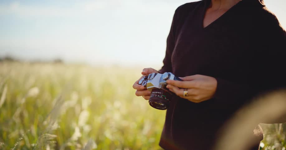 A photographer makes adjustments to her vintage film camera while standing in a sun-drenched field of tall grass