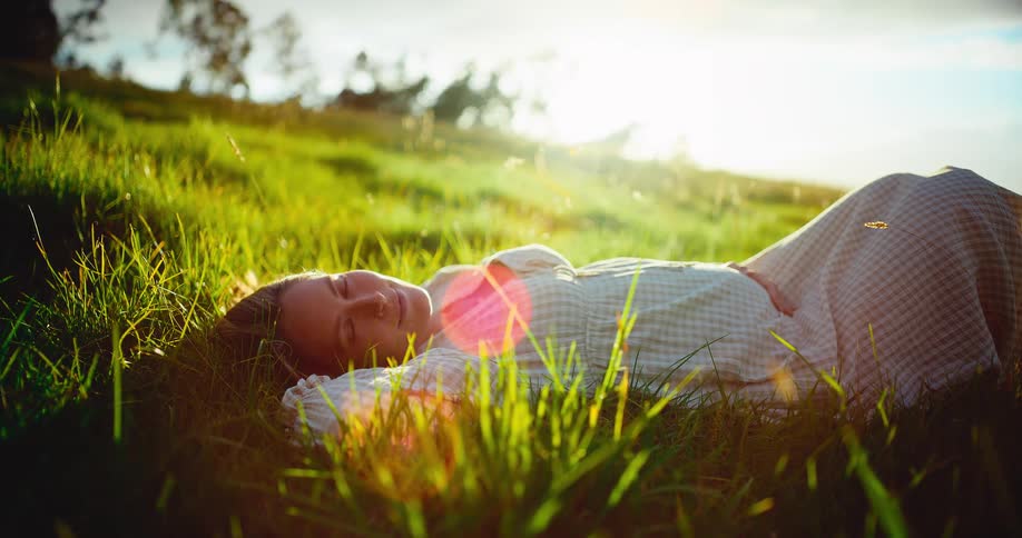 A peaceful pregnant woman relaxes in a sun-drenched grassy field, gently holding her belly as she lies back and smiles