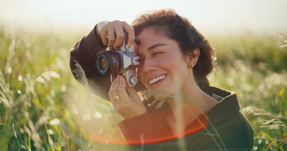A beautiful young woman smiles while taking photos with her vintage film camera in a sun-drenched field of tall grass