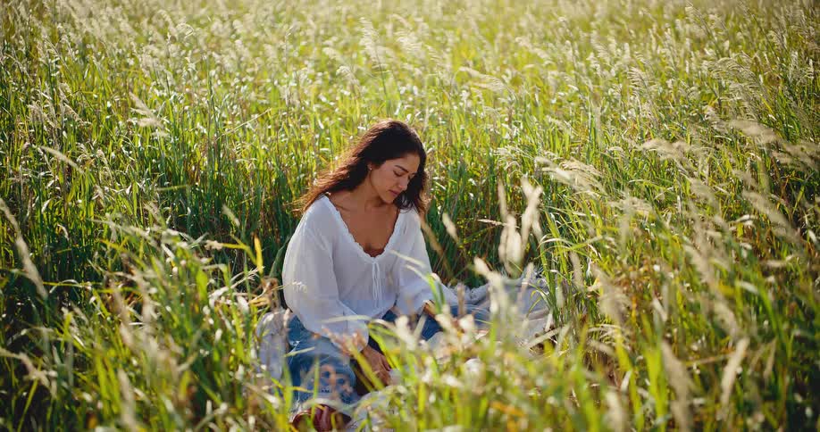 A thoughtful woman with long dark hair sits alone in a sunlit field of tall, swaying grass
