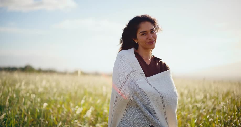 A beautiful serene woman wrapped in a cozy blanket smiles peacefully while standing in a sunlit grassy field