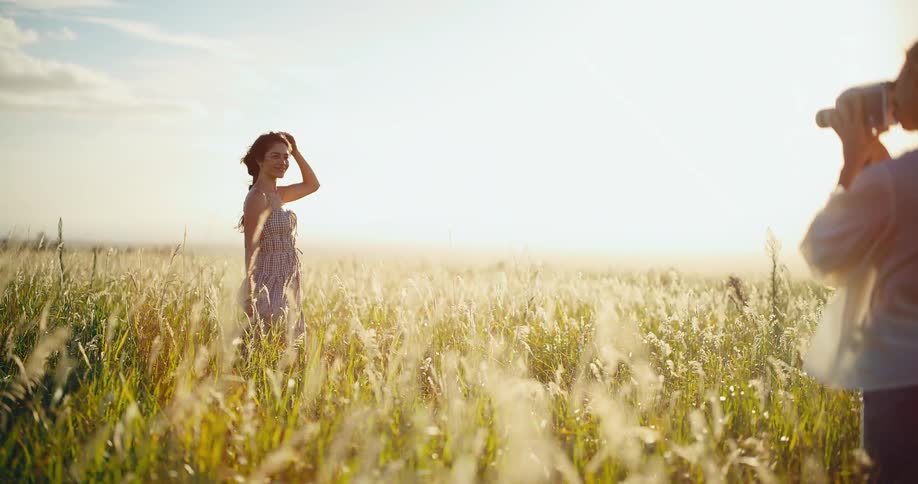 A young woman poses for a friend taking her picture with a camera in a beautiful sunlit field of tall grass at sunset
