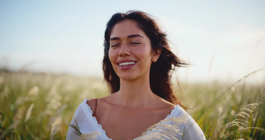 A beautiful serene woman smiles peacefully in a golden sunlit field of tall grass as the wind blows gently through her hair