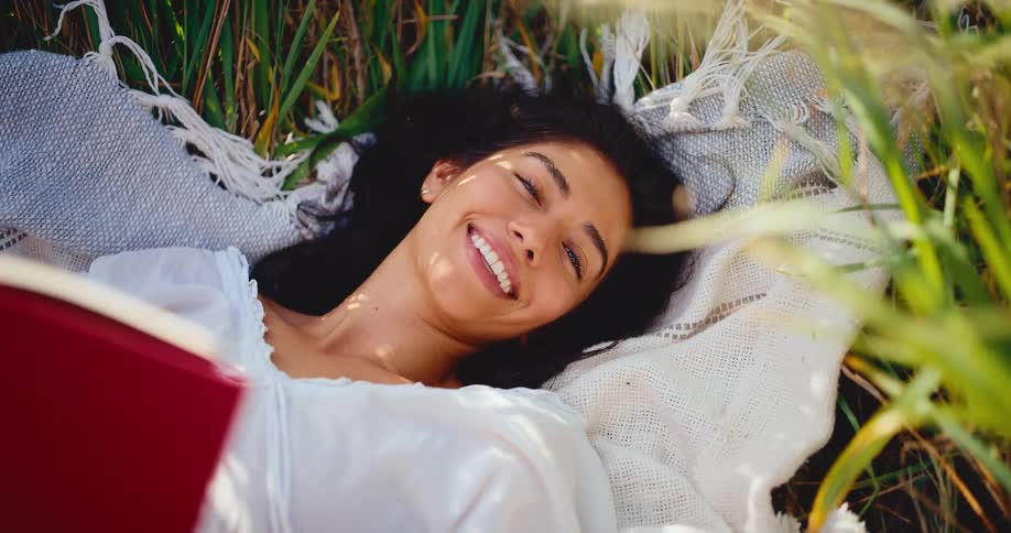 A beautiful smiling woman relaxes on a blanket in a field of tall grass while reading a book in the sun