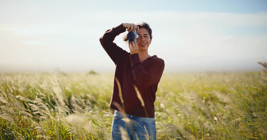 A beautiful young woman smiles and takes a picture with her vintage camera while standing in a breezy golden field in the afternoon sun