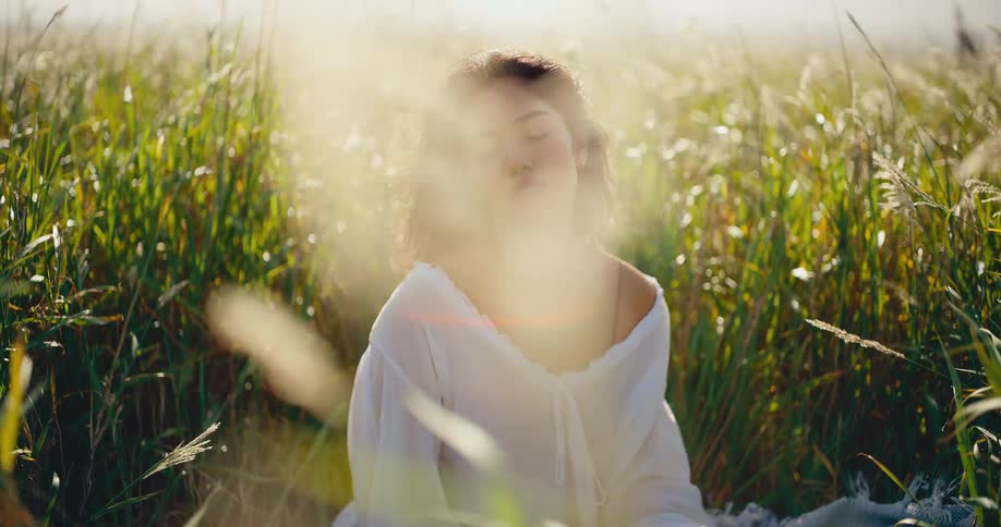 A beautiful woman sits peacefully in a sun-drenched field of tall grass, meditating with her eyes closed as the wind blows through her hair