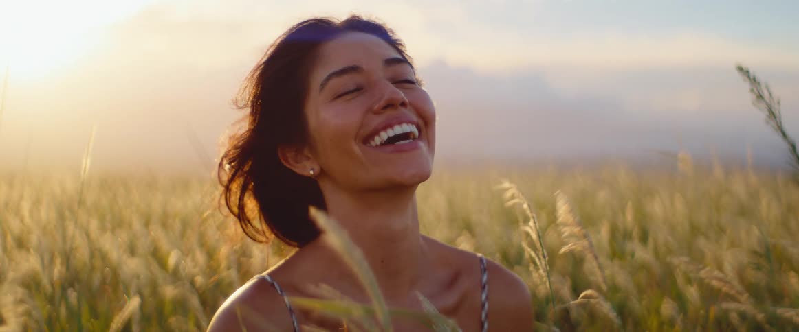 An attractive ethnic woman smiles and laughs with pure joy in a sunlit field of tall grass at sunset
