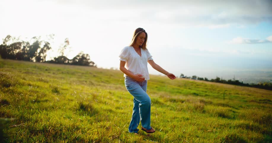 A happy pregnant woman walks through a sun-kissed green field while gently holding her belly in slow motion