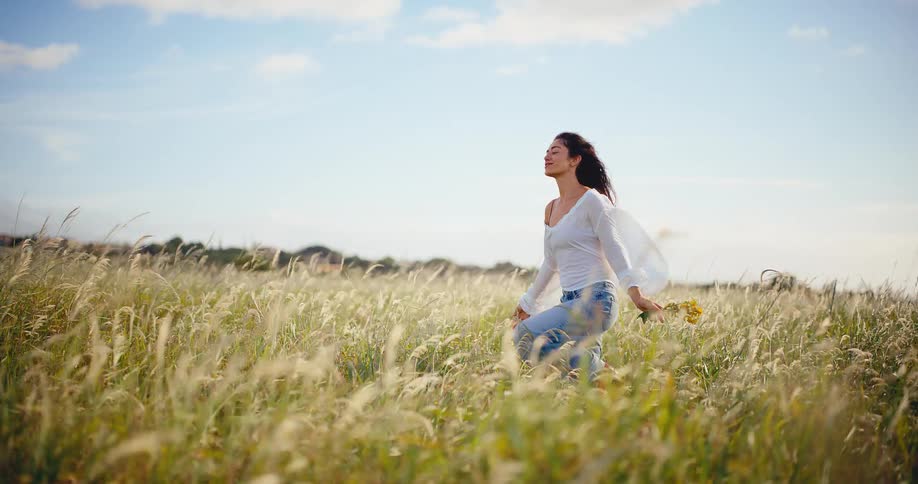Slow motion of a peaceful woman holding wildflowers while walking through a beautiful sunlit field of tall grass