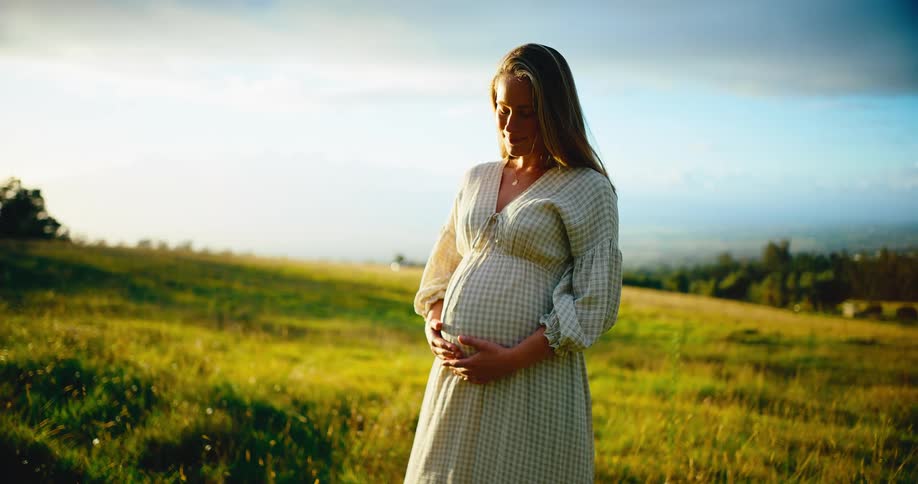 A peaceful pregnant woman lovingly holds her belly while standing in a beautiful green field at sunset