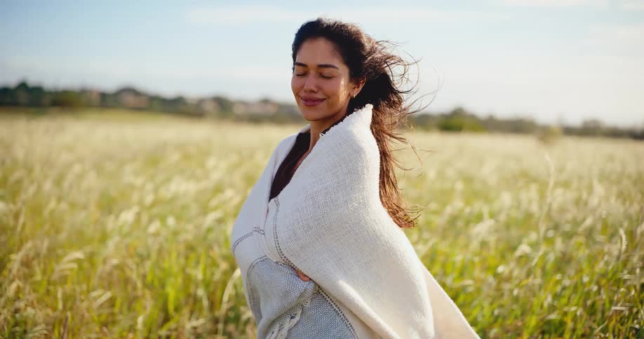 A beautiful woman wrapped in a cozy blanket smiles peacefully while standing in a sunlit field with her hair blowing in the wind