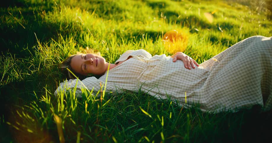 An expectant mother in a checkered dress lies peacefully in a sun-drenched green meadow, smiling as she looks up at the sky