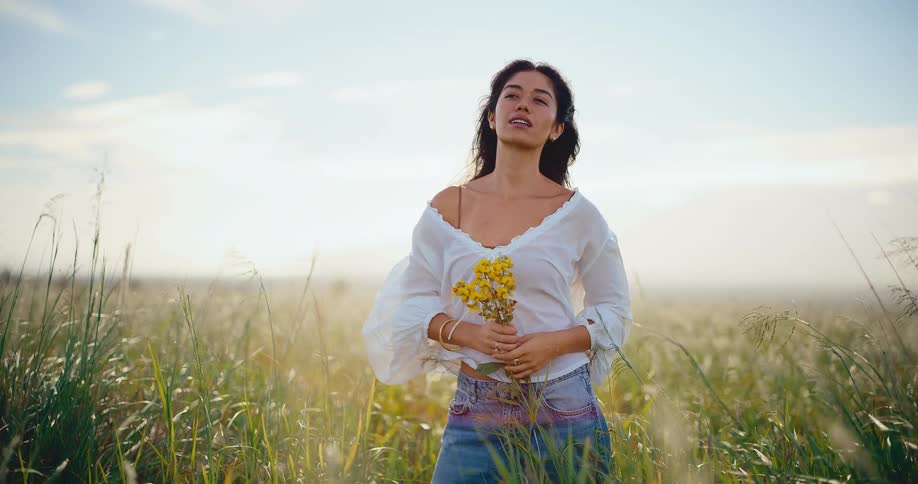 An attractive bohemian woman holding yellow wildflowers peacefully enjoys the golden hour sunlight in a windy field