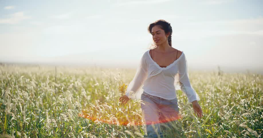 Serene young woman walks peacefully through a golden field of tall grass at sunset, holding wildflowers