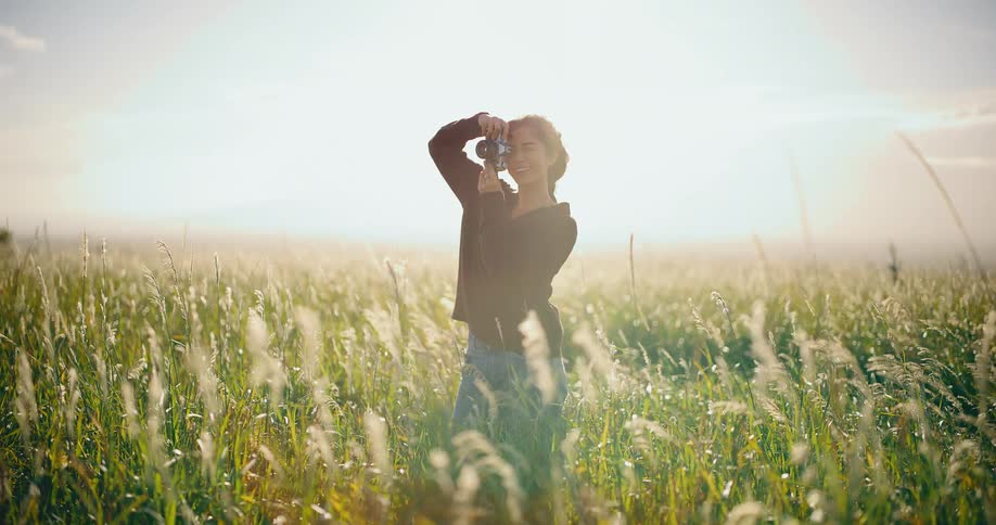 A smiling female photographer stands in a sun-drenched field of tall grass, capturing the moment with her vintage camera in slow motion