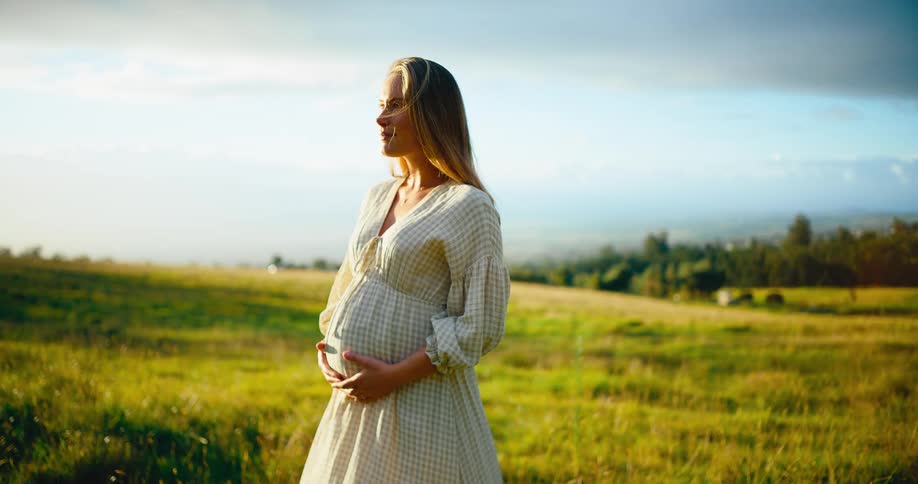 A beautiful pregnant woman gently holds her belly and smiles while standing in a grassy field, bathed in the warm light of the setting sun