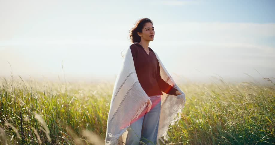 A serene woman wrapped in a blanket stands peacefully in a sun-drenched field of tall grass as the wind blows gently through her hair