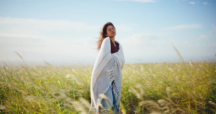 A beautiful young woman wrapped in a cozy blanket stands peacefully in a golden field of tall grass, with her hair blowing in the wind