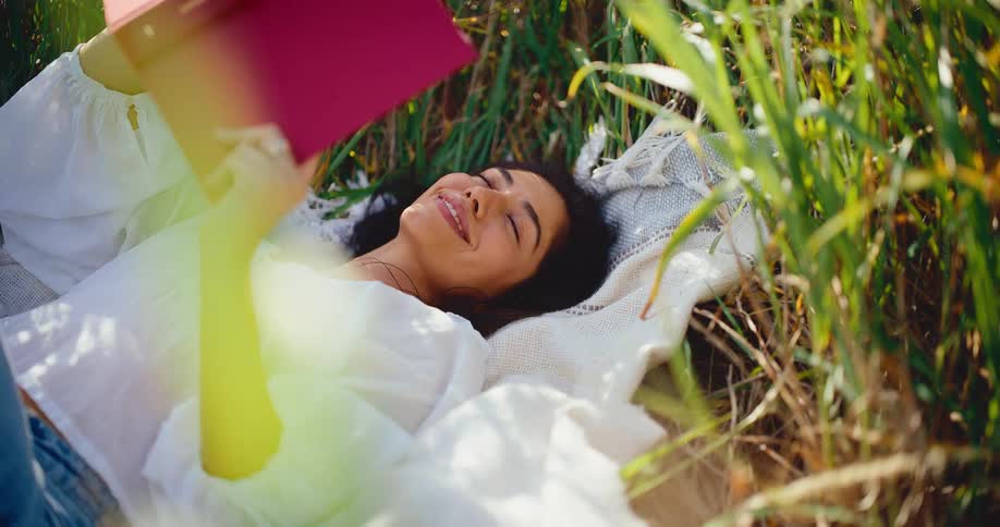A relaxed young woman smiles while reading a book, lying on a blanket in a sunlit field of tall green grass
