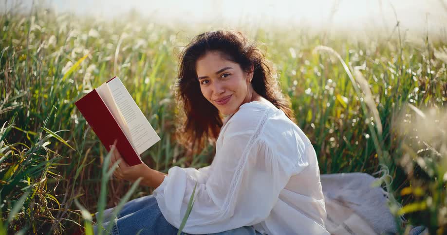 A beautiful young woman enjoys reading a book in a picturesque field of tall grass, smiling warmly at the camera in the golden sunlight