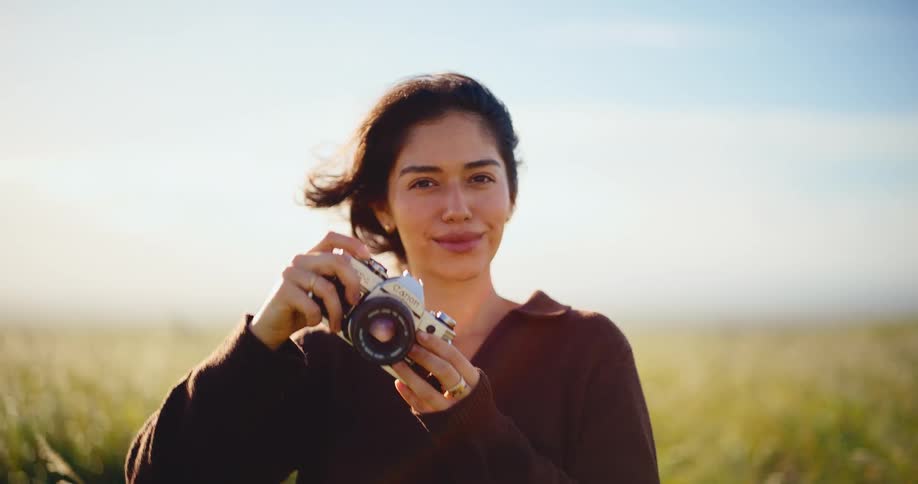 Beautiful young female photographer smiles while taking a picture with a vintage camera in a sunlit, grassy field