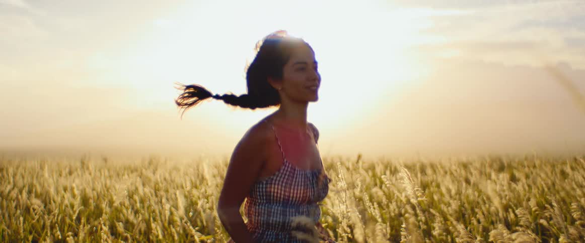 A beautiful young woman in a gingham dress runs through a sun-drenched field of tall grass at sunset in slow motion