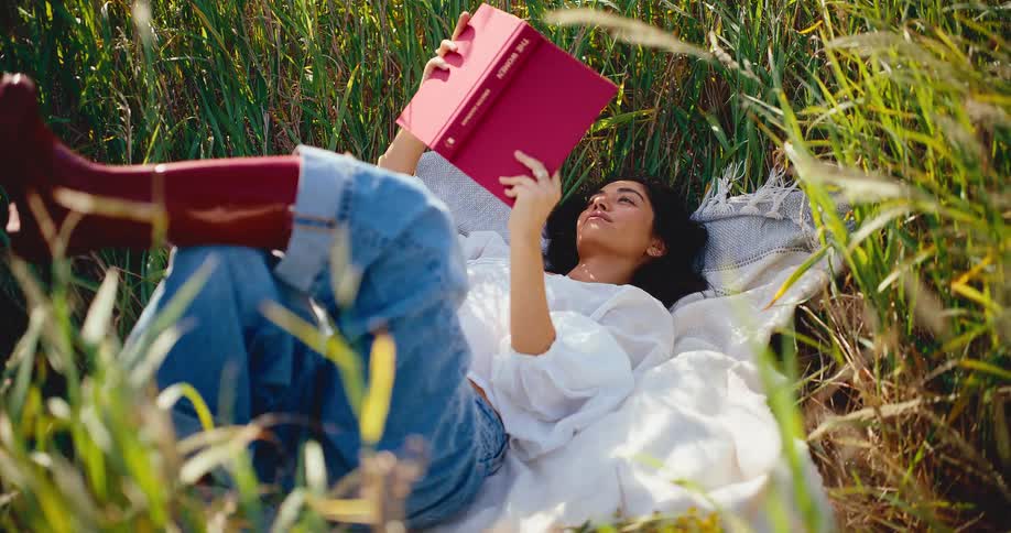 A serene woman in red boots relaxes on a blanket in a tall grass field, enjoying a book in the sunshine