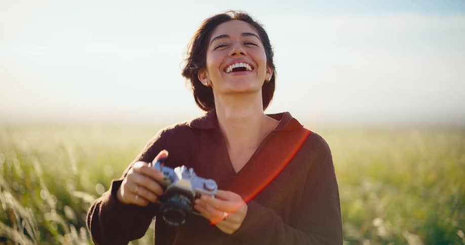 Beautiful young woman laughs while taking pictures with her vintage camera in a golden field at sunset