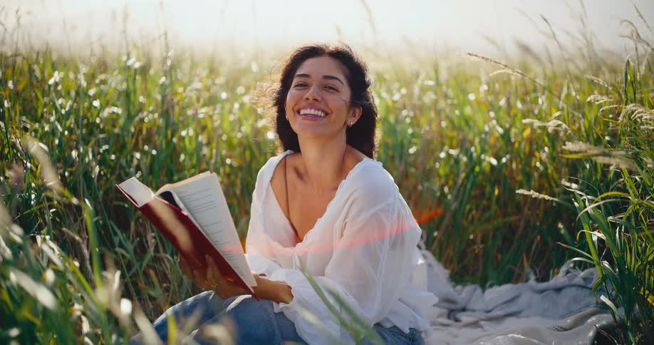 A beautiful woman in a white blouse reads a book and smiles peacefully while sitting on a blanket in a breezy, sun-drenched field