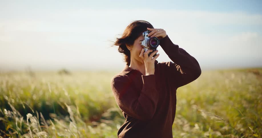 A joyful woman with a vintage camera laughs and takes pictures while standing in a sun-drenched field of tall grass blowing in the wind