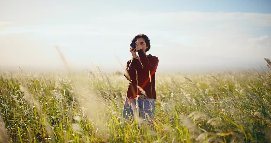 Beautiful young woman taking pictures with a vintage camera in a golden field of tall grass at sunset