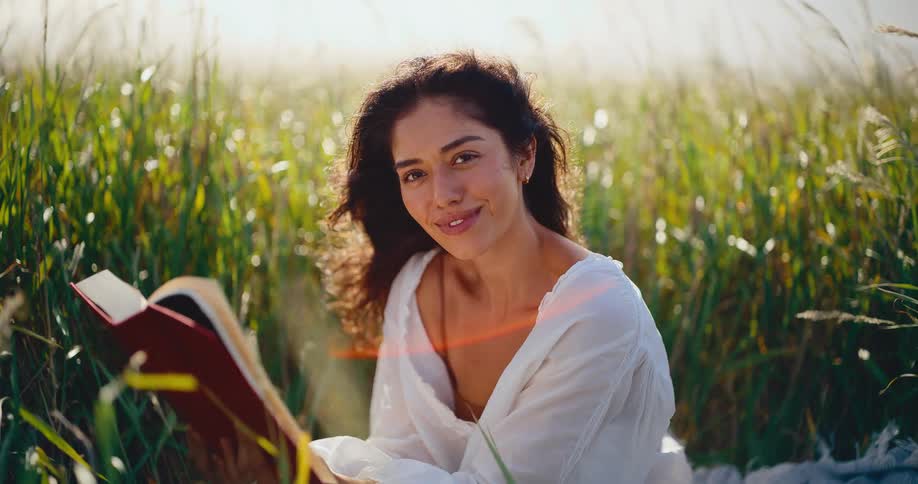 Portrait of a happy young woman laughing and smiling while reading a book in a sunny meadow in slow motion