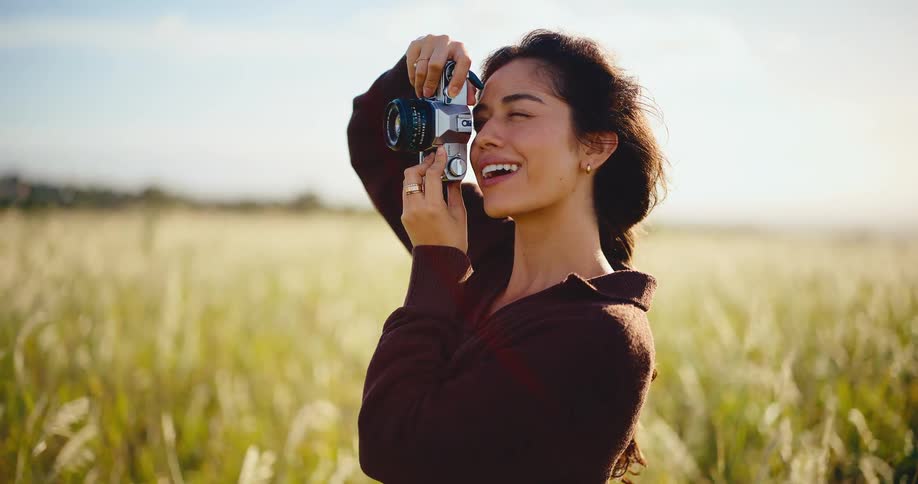A smiling female photographer captures a moment with her retro film camera in a sun-drenched field of tall grass