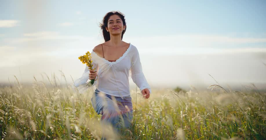A beautiful young woman walks through a sunlit field of tall grass, smiling and smelling a bouquet of wildflowers in slow motion
