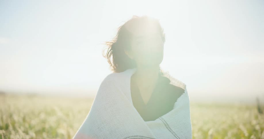 A serene young woman wrapped in a blanket smiles and soaks in the warm sun while standing in a breezy, golden field