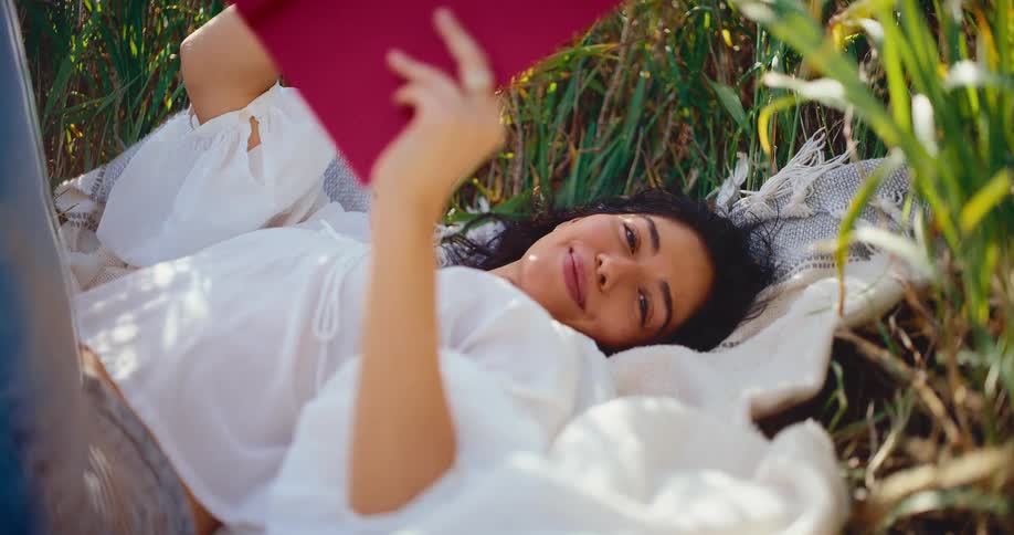 A beautiful woman smiles contentedly while lying on a blanket in a sun-drenched grassy field, enjoying a book in slow motion