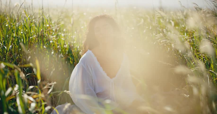 A beautiful serene woman sits peacefully in a golden sunlit field of tall grass, closing her eyes to feel the moment