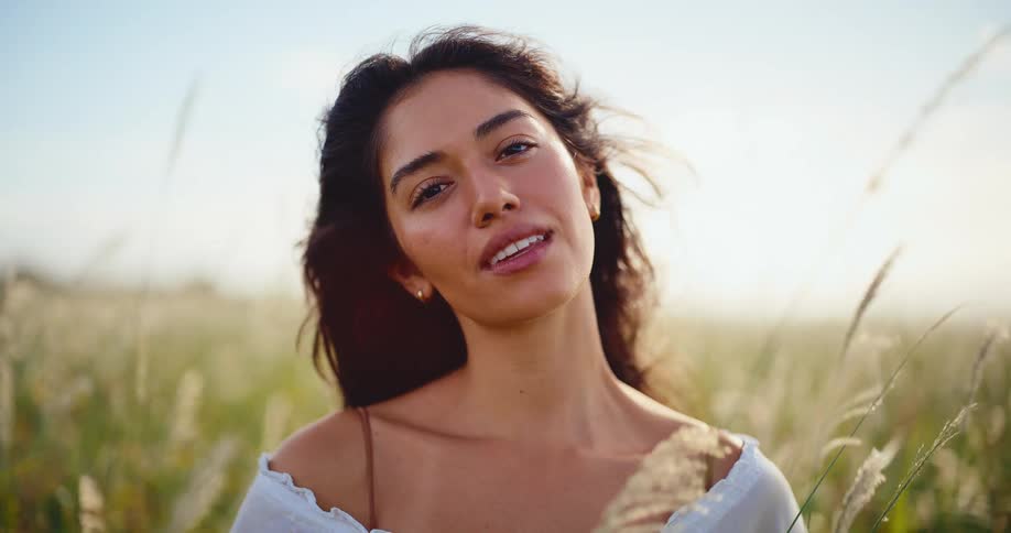A beautiful young woman smiles peacefully at the camera while standing in a breezy field of tall grass during a golden sunset