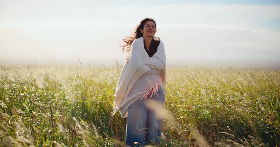 Beautiful woman wrapped in a warm blanket enjoys the afternoon sun in a windy golden field in slow motion