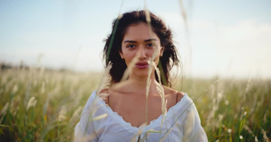 A serene young woman in a white blouse closes her eyes and smiles peacefully, enjoying the warm sunlight in a beautiful field of tall grass