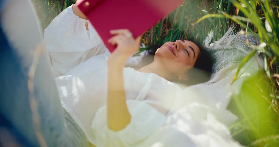A serene young woman peacefully reads a book while lying on a blanket in a sun-drenched grassy field