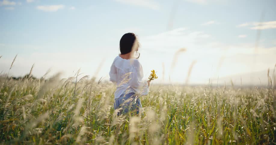 A tranquil scene from behind of a woman holding wildflowers while standing in a sun-drenched, breezy field of tall grass